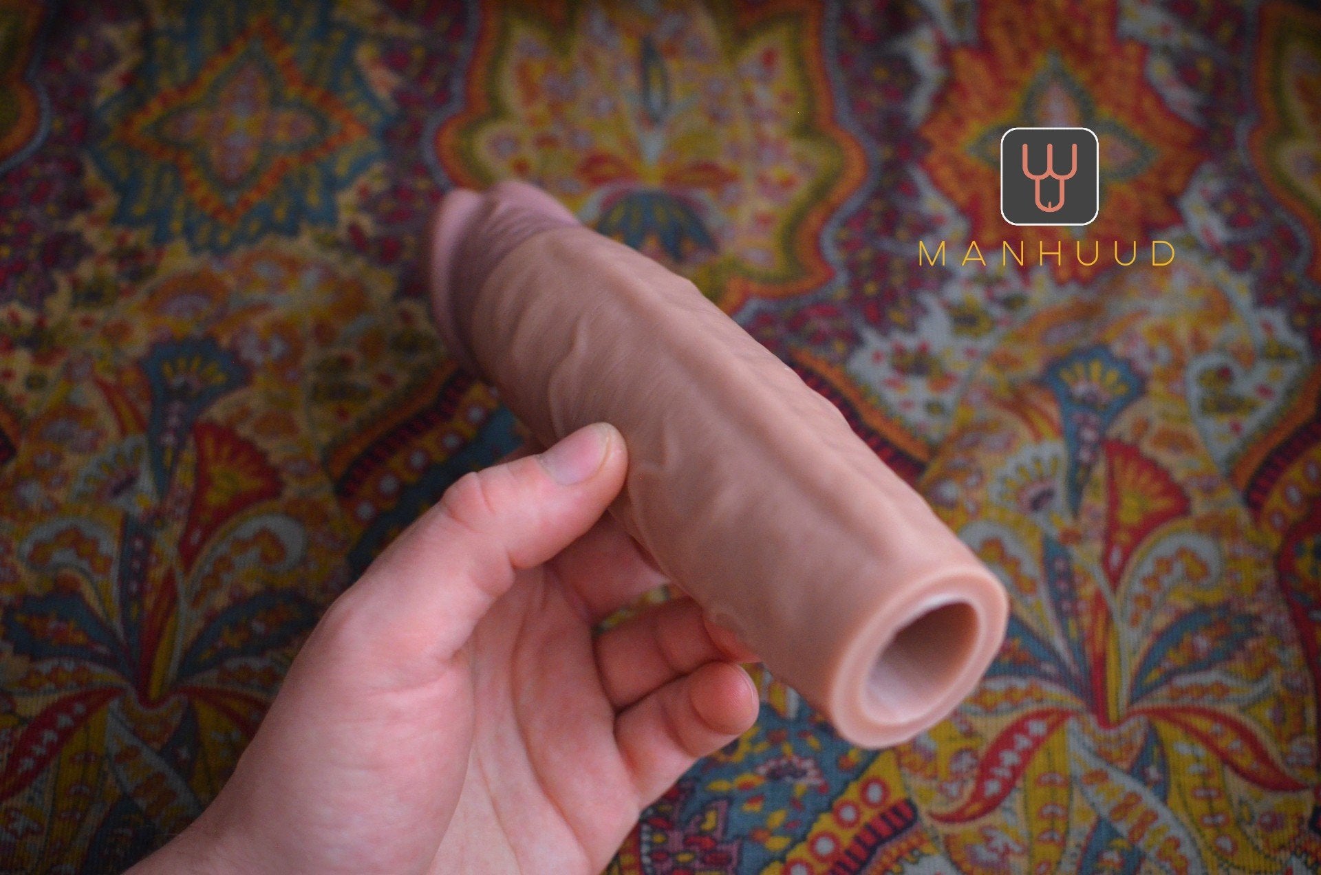 Hand holding a realistic silicone sleeve with an open end against a colorful patterned background, labeled "MANHUUD"