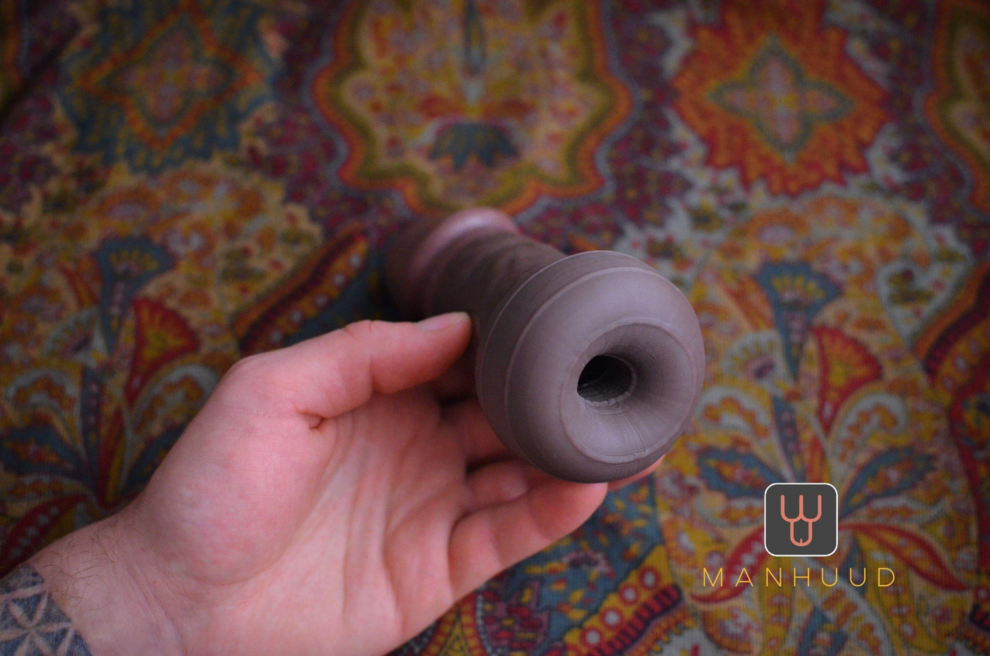 Hand holding a brown silicone model with an open end against a colorful patterned background, labeled "MANHUUD"